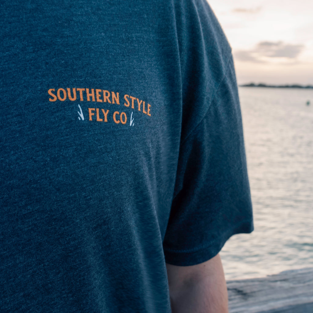 Close-up macro of the front chest logo on a charcoal Southern Style Fly t-shirt, shot on a dock with blurred water in the background, highlighting fabric texture and clean branding.