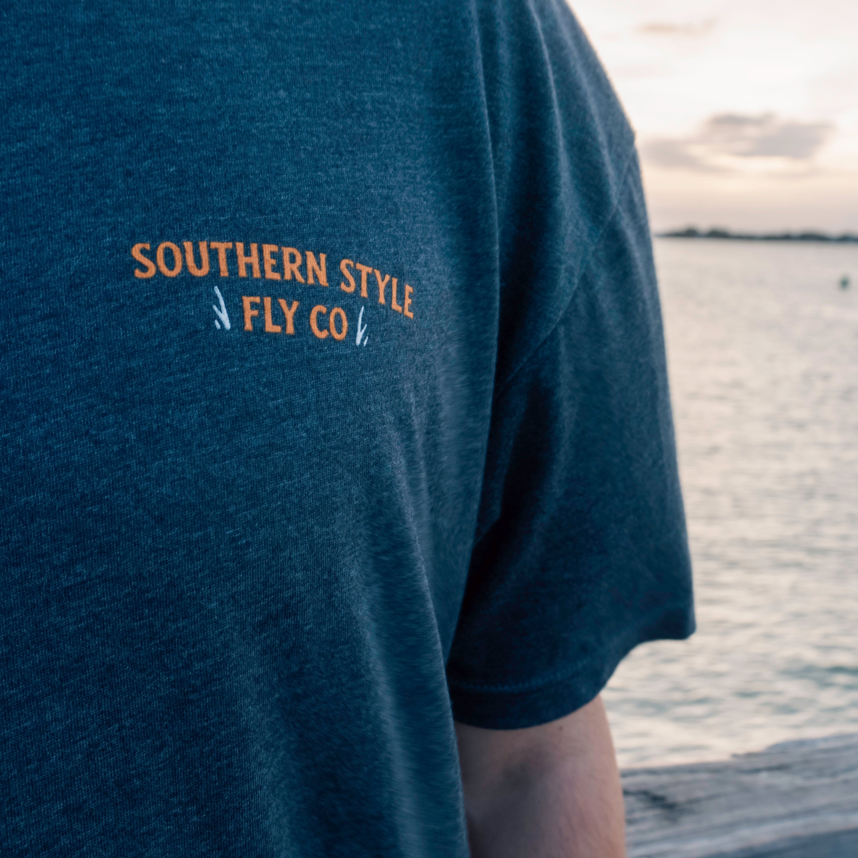 Close-up macro of the front chest logo on a charcoal Southern Style Fly t-shirt, shot on a dock with blurred water in the background, highlighting fabric texture and clean branding.