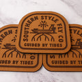 Tan 3.7 x 4 inch Southern Style Fly Co. peel-and-stick foam fly pad featuring the Guided By Tides Skiff logo with a skiff and mangrove design, built for mounting on fly boxes, coolers, and boat consoles.