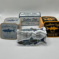 Assorted Southern Style Fly Co. fishing stickers displayed on a white background, including square logo stickers in multiple colorways and a stacked die-cut camo fish sticker in the foreground.