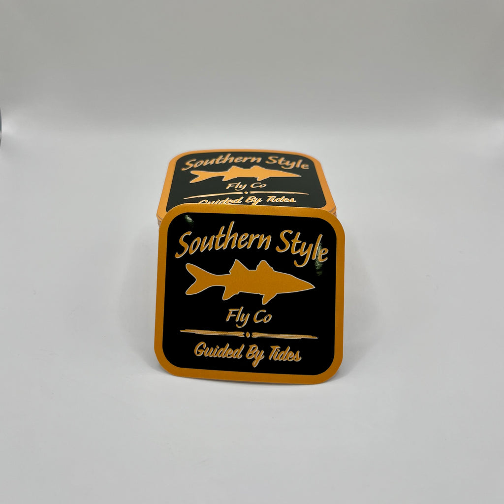 Macro close-up of a Southern Style Fly Co. square sticker with a black background, gold lettering, and a gold fish logo.