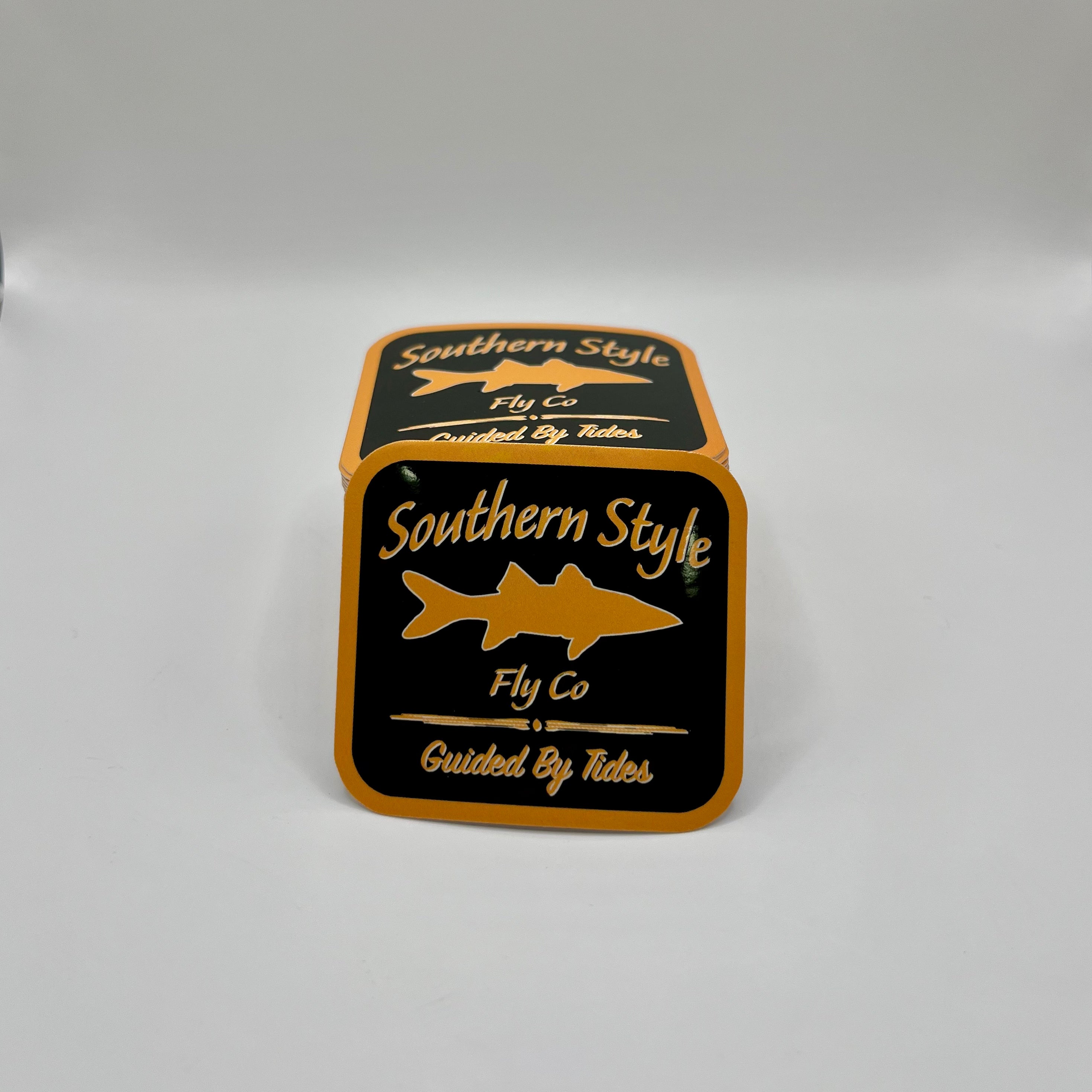 Macro close-up of a Southern Style Fly Co. square sticker with a black background, gold lettering, and a gold fish logo.
