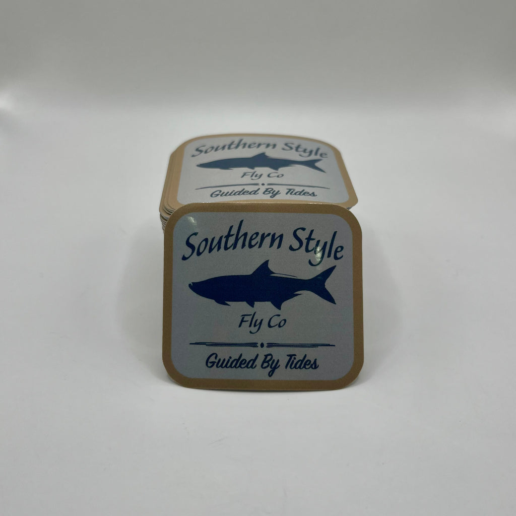 Macro close-up of a Southern Style Fly Co. square sticker with a tan background, blue fish logo, and “Guided by Tides” text.
