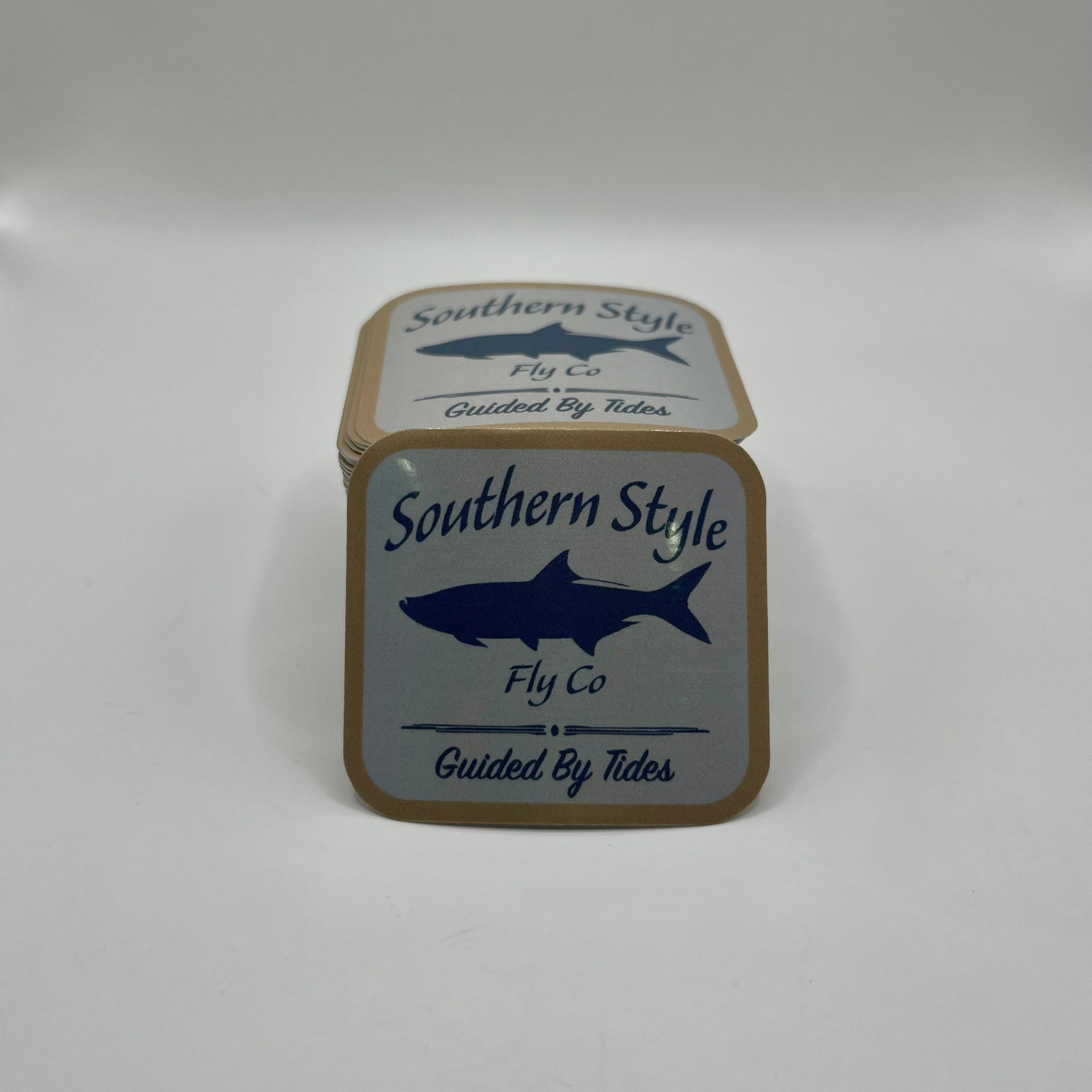 Macro close-up of a Southern Style Fly Co. square sticker with a tan background, blue fish logo, and “Guided by Tides” text.