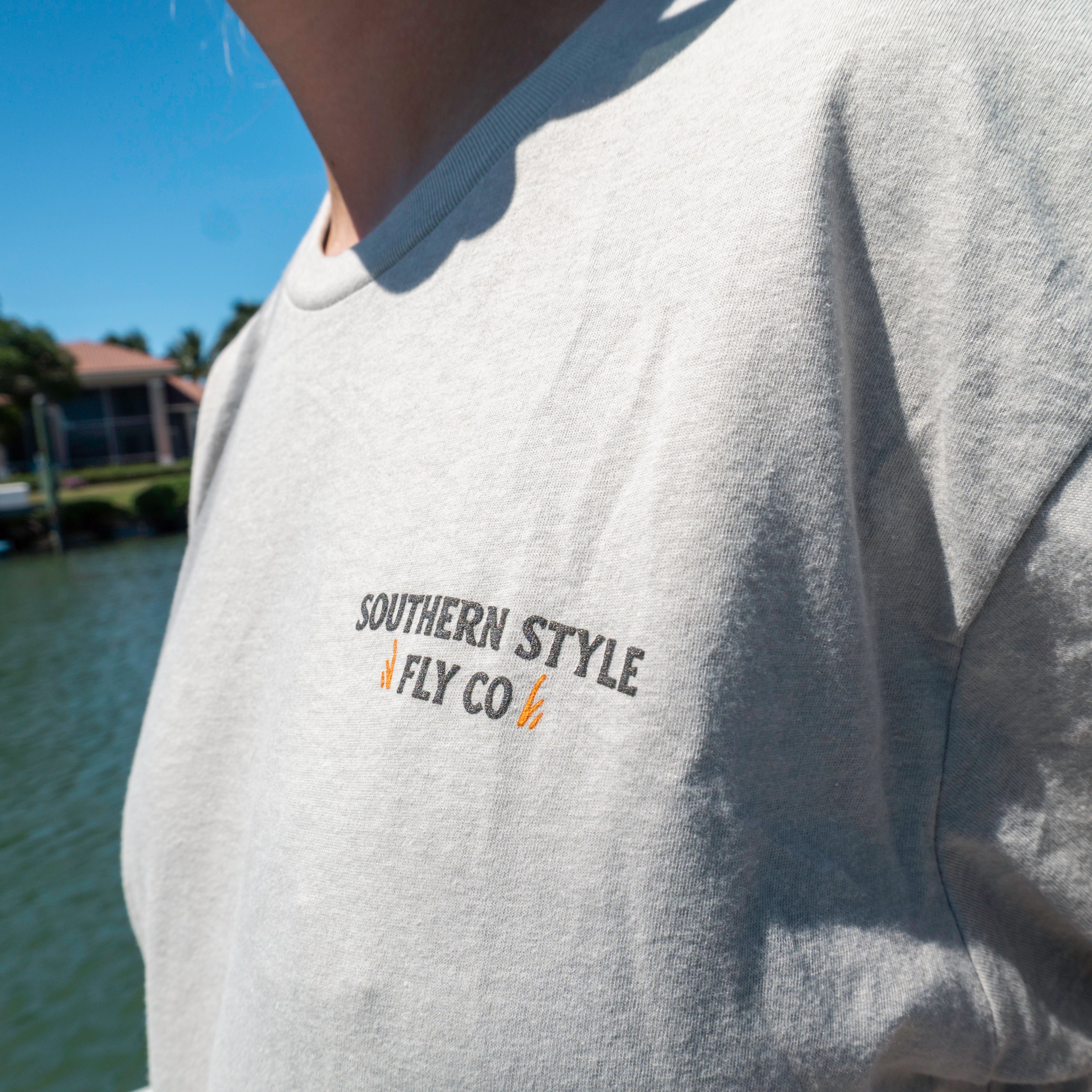 Close-up macro of the front chest logo on a silk Southern Style Fly t-shirt, shot on a boat with blurred water in the background, highlighting fabric texture and clean branding.