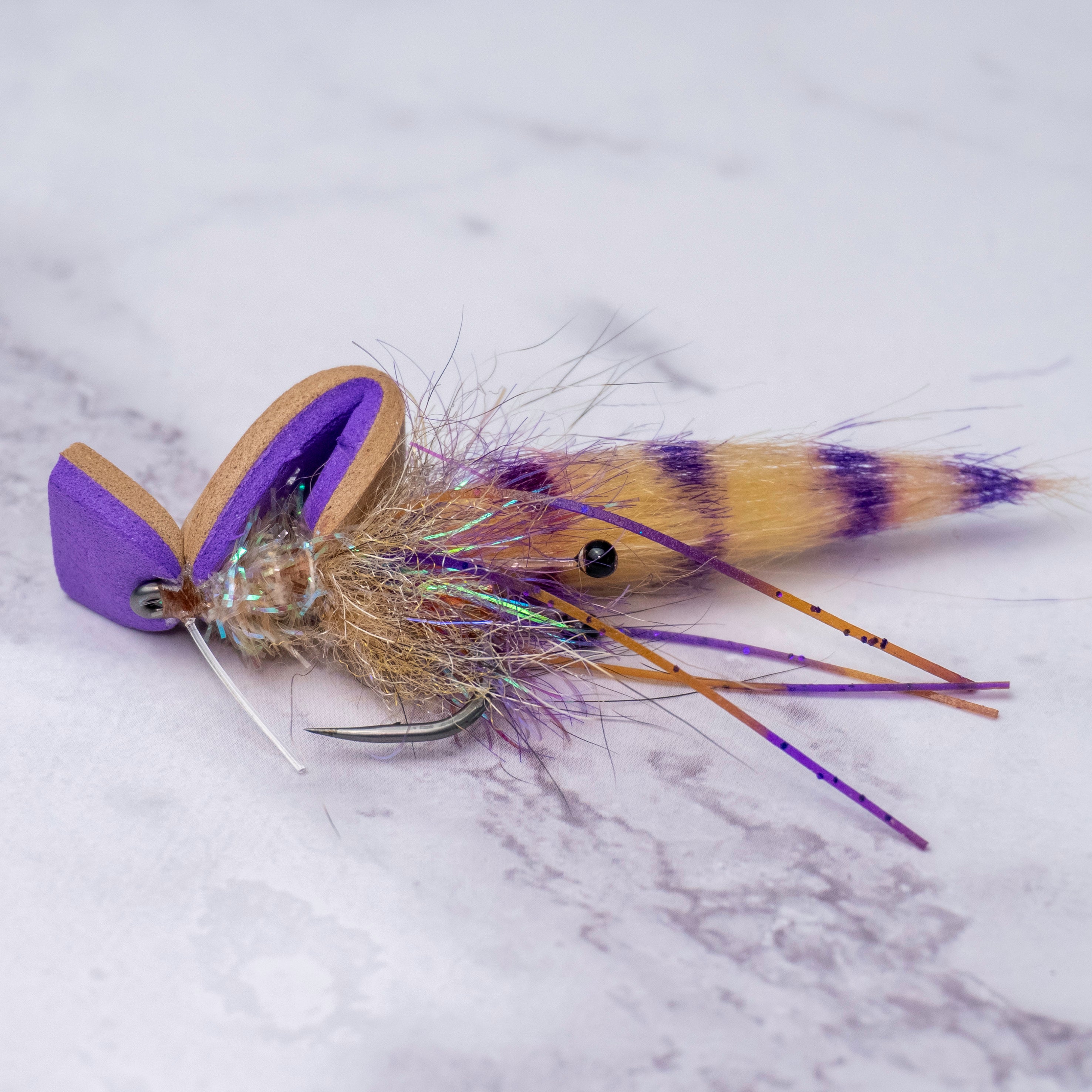 Close-up of the limited batch Summer Collection Pop-N-Shrimp fly featuring purple foam popping head, barred tail, flash body, rubber legs, and premium saltwater hook on white marble surface.