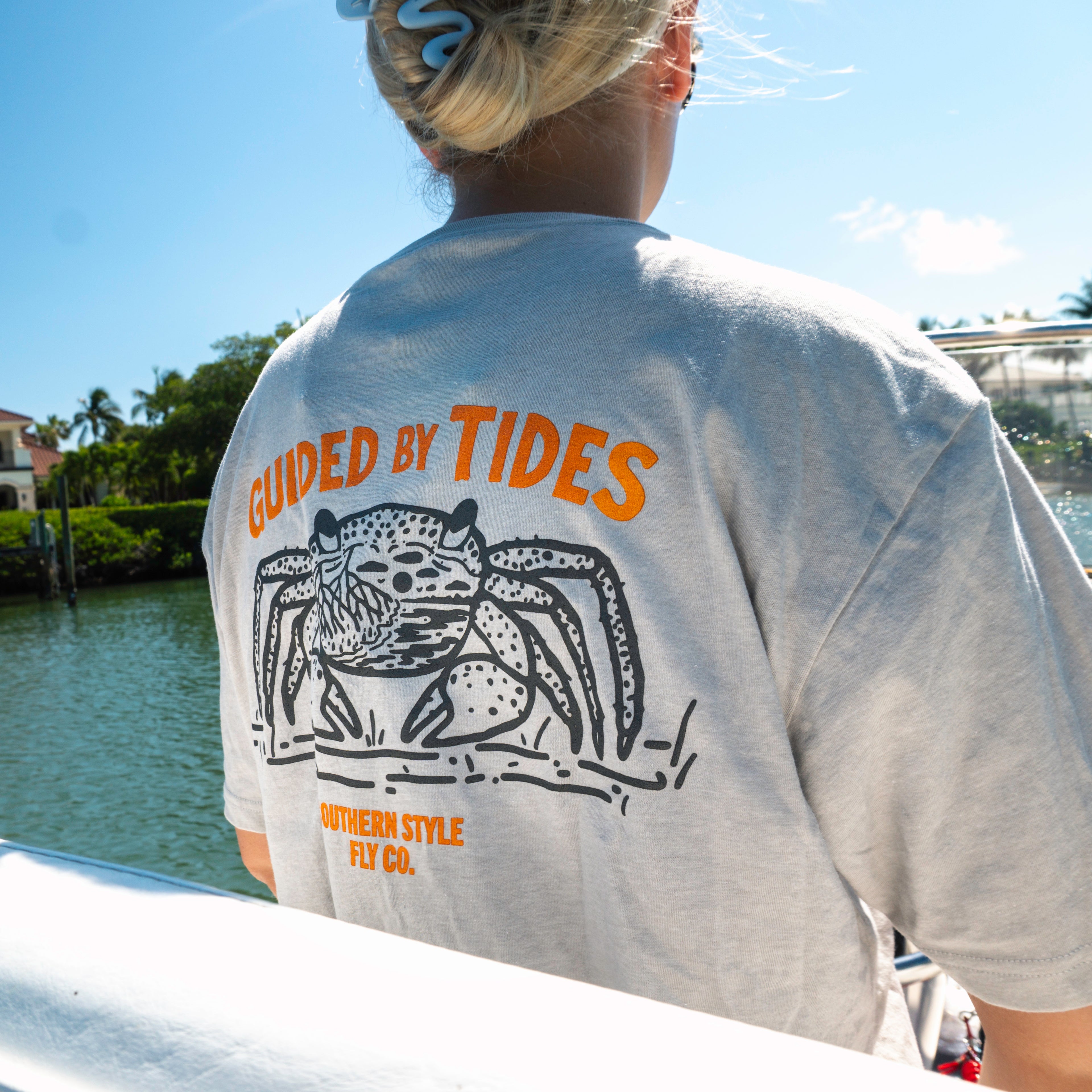 Angler wearing black “Guided by Tides” Southern Style Fly t-shirt while driving a boat, capturing an on-the-water summer fishing lifestyle scene with focus on movement, sunlit conditions, and active boating environment.