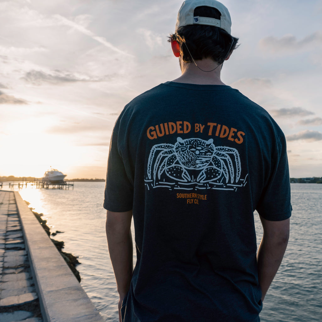 Person wearing charcoal “Guided by Tides” t-shirt viewed from the back, standing on a seawall and looking out over the water at sunset, showcasing the large crab graphic in a coastal lifestyle setting.