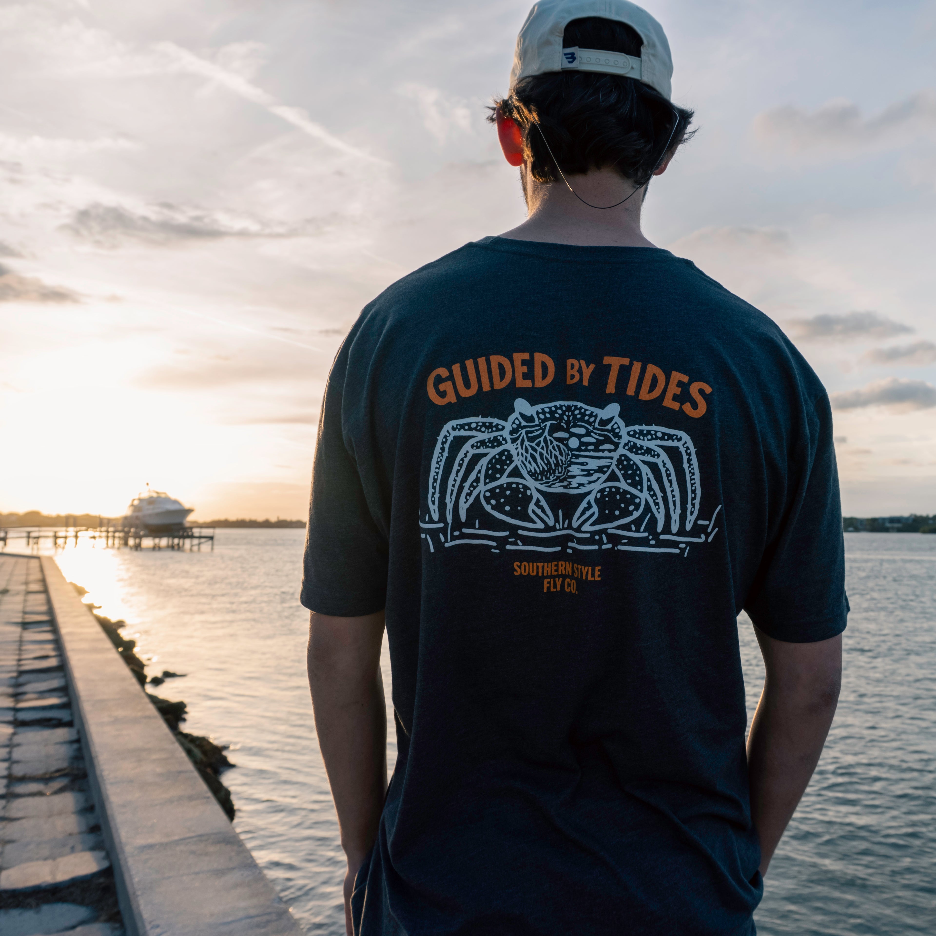 Person wearing charcoal “Guided by Tides” t-shirt viewed from the back, standing on a seawall and looking out over the water at sunset, showcasing the large crab graphic in a coastal lifestyle setting.