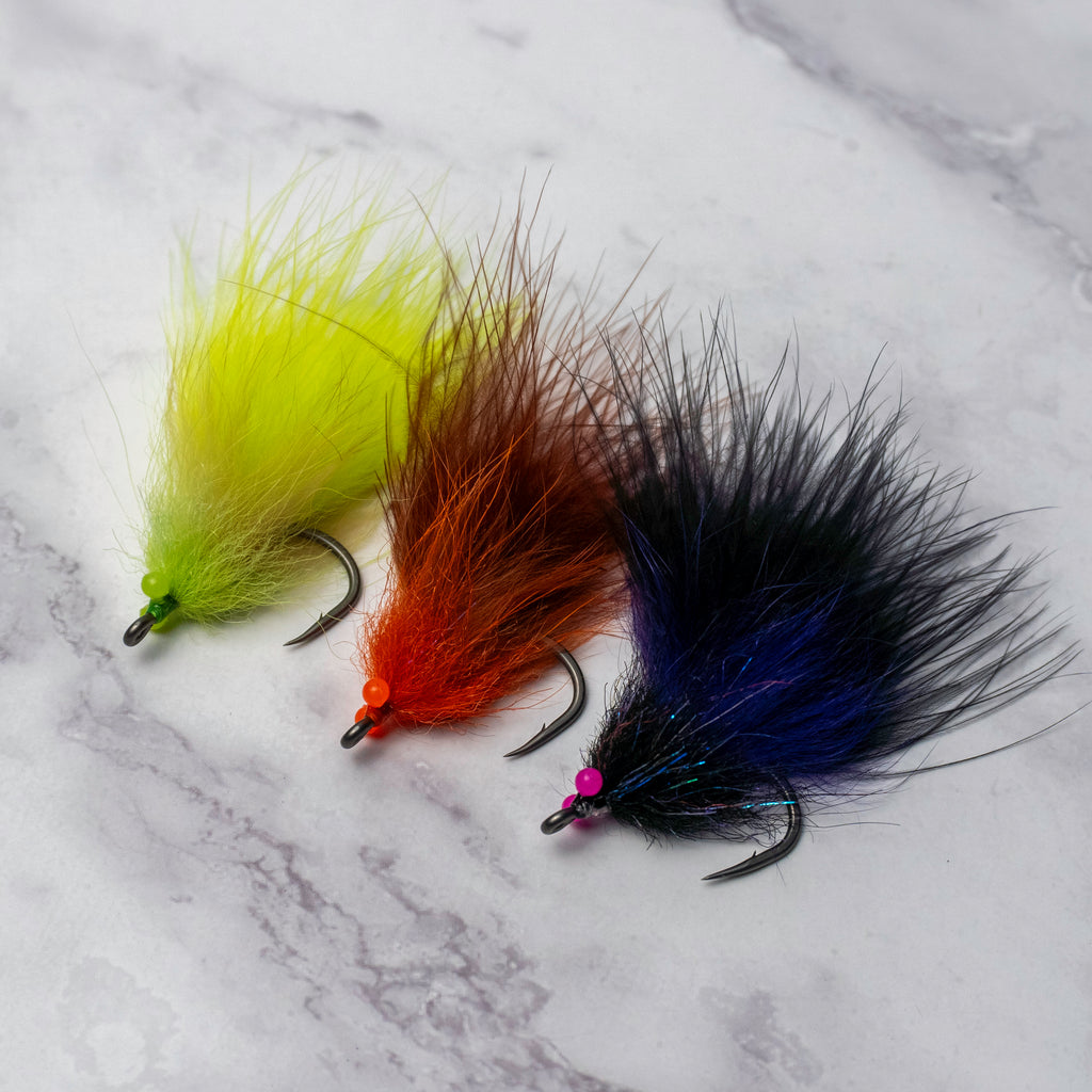 Three South Bounder tarpon fly color variations including chartreuse, orange brown, and black purple displayed together on a light marble background.