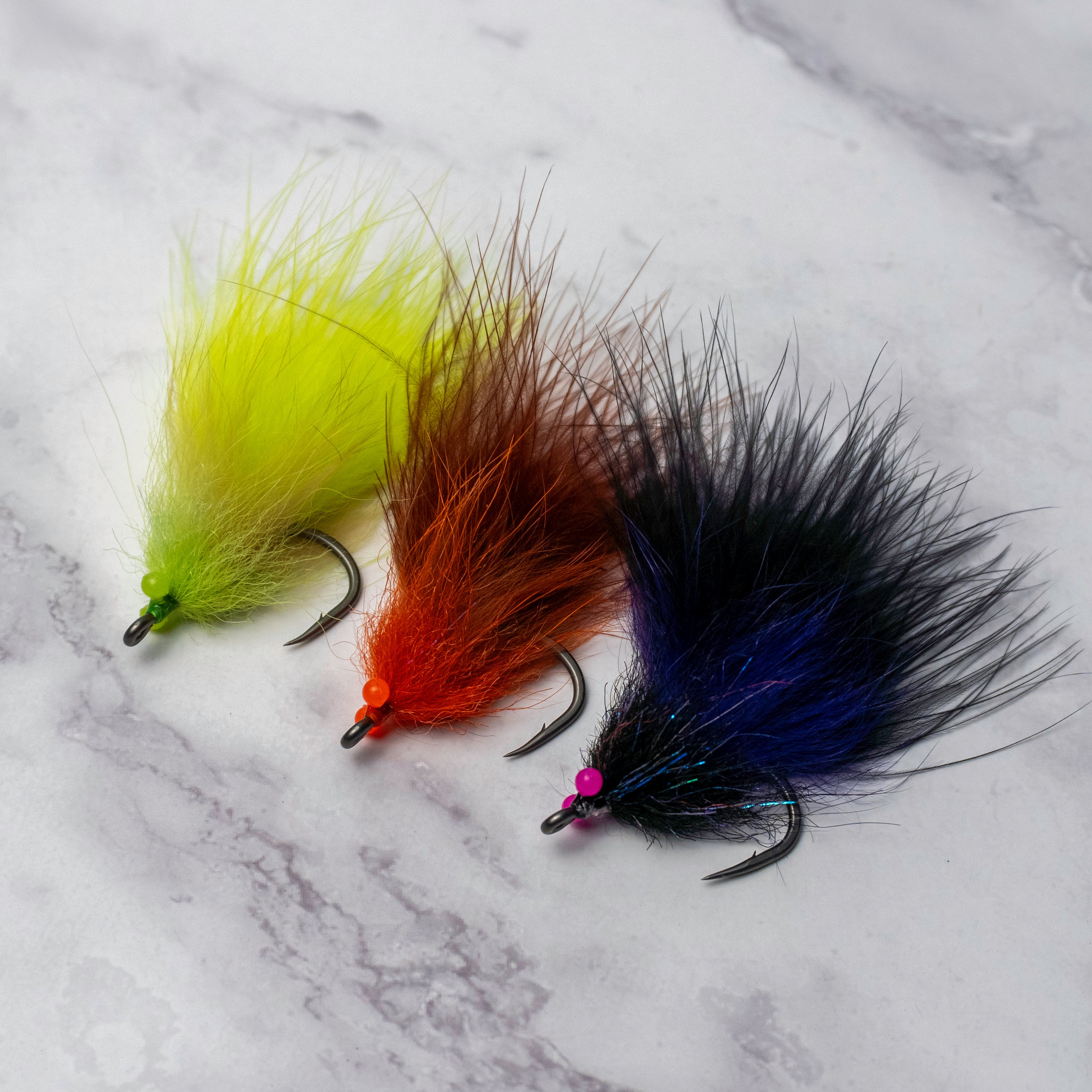 Three South Bounder tarpon fly color variations including chartreuse, orange brown, and black purple displayed together on a light marble background.