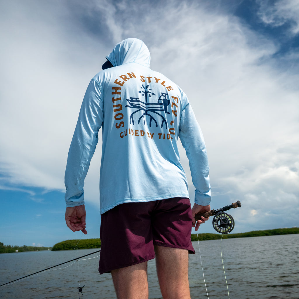 Fly angler wearing the light blue Tidebound Tech Hoodie and holding a fly rod beside a mangrove shoreline